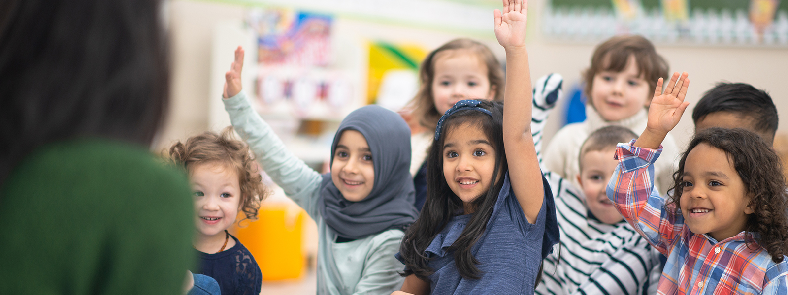 A multi-ethnic group of children in their classroom. They are sitting on the floor and raising their hand to answer a question posed by their teacher while reading a story. They are all smiling.